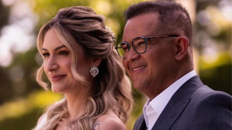 Livia e Bruno Avelar em clima romântico ao ar livre, com Lívia de cabelo ondulado e brincos brilhantes e Bruno Avelar usando óculos e terno, sorrindo em um cenário desfocado.
