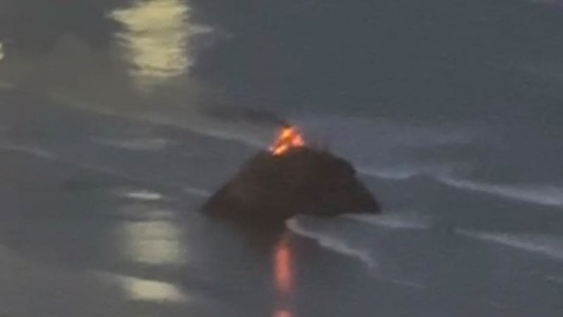 Cena desfocada com fumaça e luzes ao fundo, sugerindo fogo ou incêndio em meio a um cenário noturno, com clarões refletidos no ambiente
