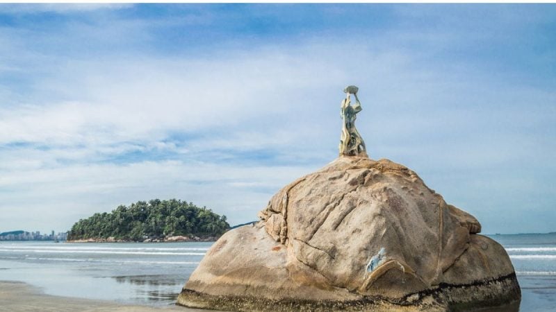 Estátua Pedra da Feiticeira em uma rocha na praia, com o mar ao fundo e céu azul ao entardecer, criando uma paisagem costeira tranquila.