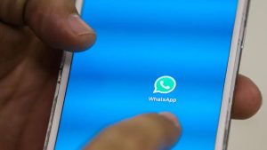 Smartphone com o aplicativo WhatsApp aberto na tela azul, com o ícone do WhatsApp central e mãos segurando o celular durante o uso