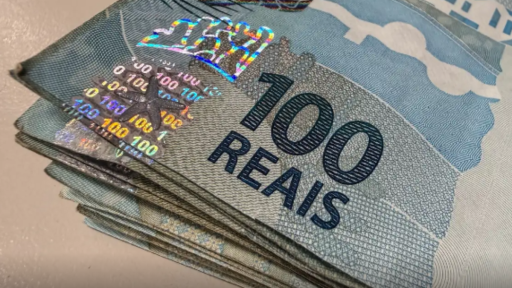 Cédulas de 100 reais com a palavra “100 REAIS” visível, imagem de dinheiro brasileiro sobre fundo claro, sugerindo dinheiro e economia.