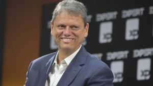 Retrato do Tarcísio de Freitas sorrindo em evento, usando terno azul e camisa branca, com fundo desfocado de painel com marcações e números.