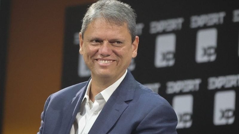 Retrato do Tarcísio de Freitas sorrindo em evento, usando terno azul e camisa branca, com fundo desfocado de painel com marcações e números.