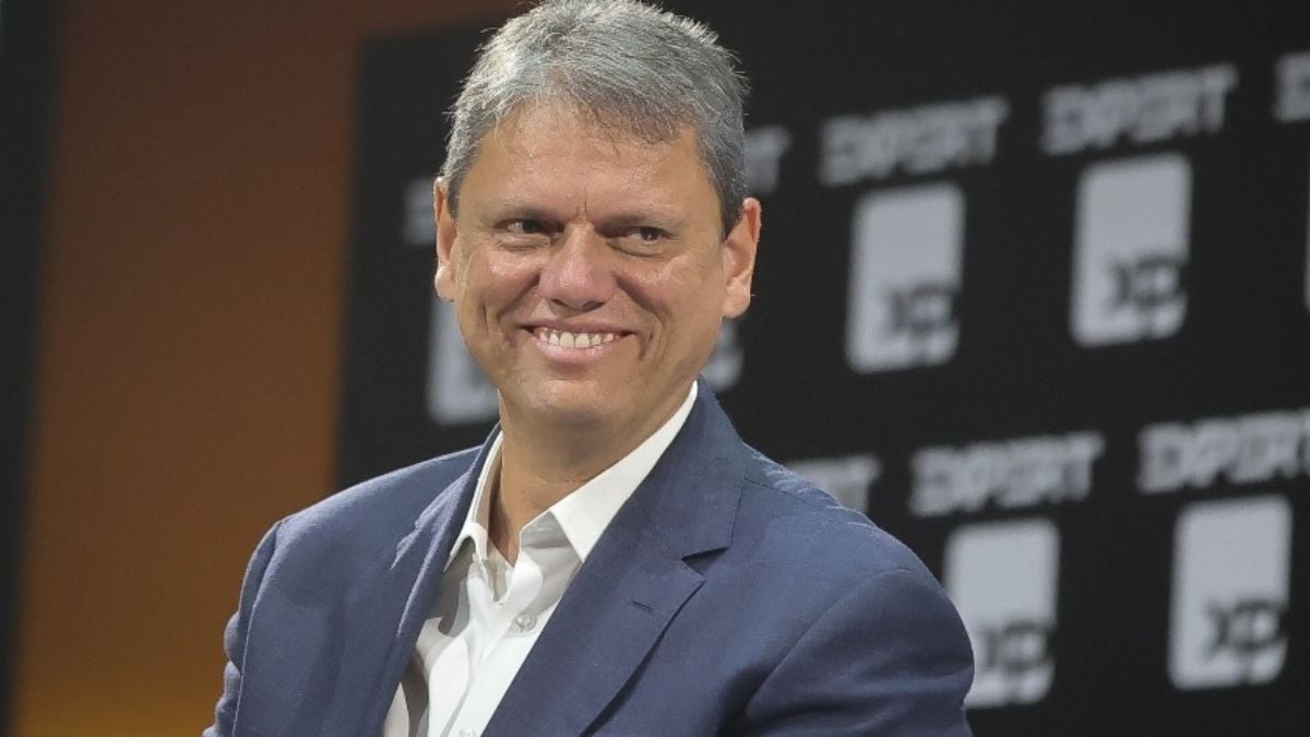 Retrato do Tarcísio de Freitas sorrindo em evento, usando terno azul e camisa branca, com fundo desfocado de painel com marcações e números.