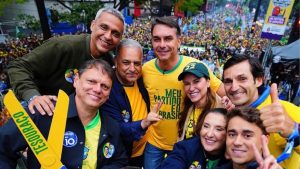 Grupo de apoiadores de Flávio Bolsonaro posa para foto durante manifestação com bandeiras do Brasil e grande público ao fundo.