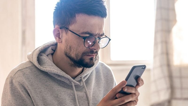 Homem usando o celular, com expressão concentrada, sentado em ambiente interno e iluminado, representando uma pessoa acompanhando informações financeiras pelo smartphone.