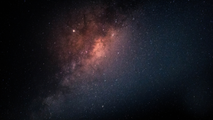 Céu noturno com estrelas e a Via Láctea em destaque, mostrando uma nebulosa luminosa em tons de rosa e vermelho contra o fundo escuro do espaço