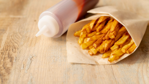 Embalagem de papel de batatas fritas crocantes temperadas em um cone, sobre bancada de madeira clara; imagem promocional de comida de rua. Ao lado uma bisnaga de molho