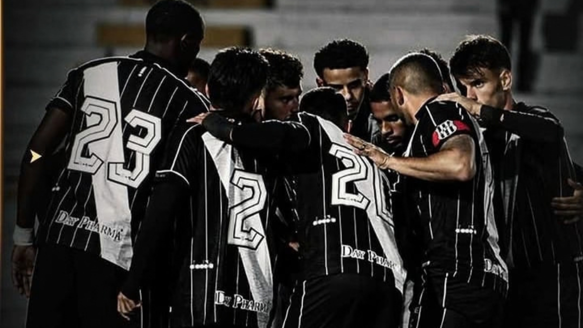 Grupo de jogadores de futebol com camisas pretas e brancas, números grandes nas costas, reunidos em meio a conversas e concentração durante um jogo ou treino em ambiente externo
