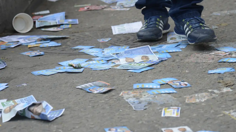 Pessoas passam próximo a um chão de rua com vários santinhos, com um par de tênis ao lado e papéis no asfalto.