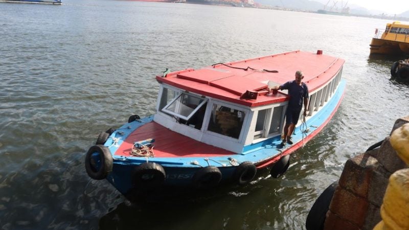 Barco de pequeno porte em cores azul, vermelho e branco. Homem se apoia na lateral do barco