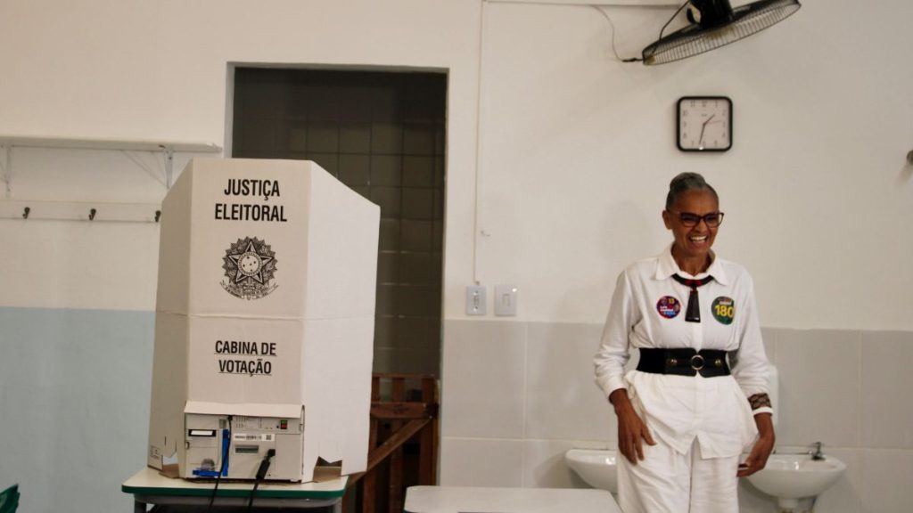 Candidata ao Senado Marina Silva aparece em foto com vestido branco e detalhes em preto, ao lado esquerdo aparece uma urna eletrônica estrito 'JUSTIÇA ELEITORAL'