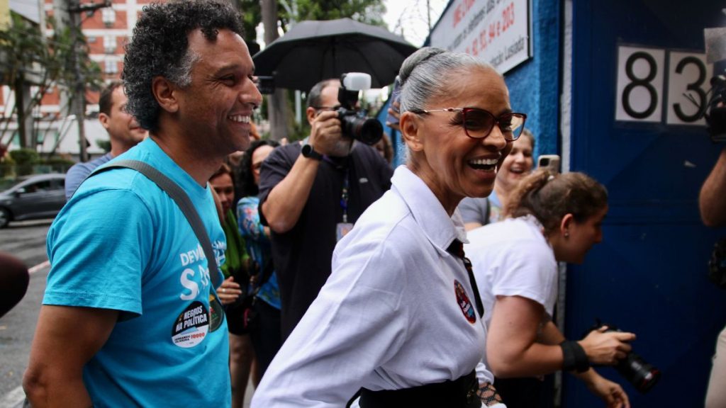 Marina Silva caminha ao lado de homem. Els esta com vestido branco e detalhes em preto, sorri ao caminhar