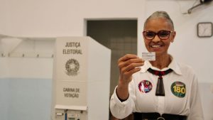 Marina Silva sorri ao mostrar o comprovante do voto nas eleições 2026. Do lado esquerdo aparece uma urna eletrônica escrito 'JUSTIÇA ELEITORAL'