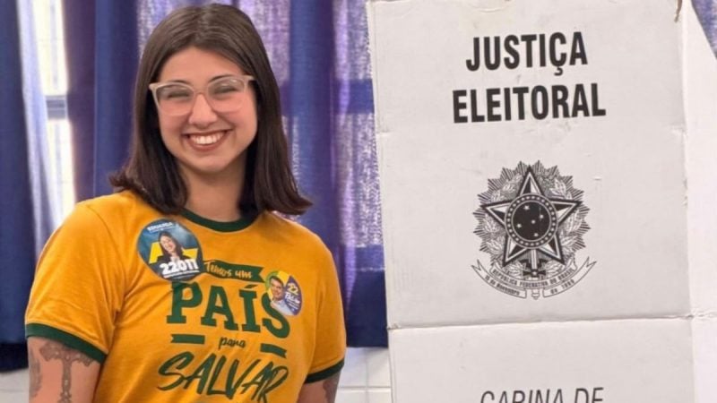 Eduarda Campopiano sorri para foto e usa uma camiseta amarela com adesivos, do seu lado direito aparece uma urna eletrônica escrito 'JUSTIÇA ELEITORAL'