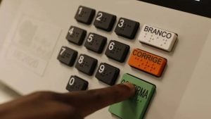Pergunta sobre segundo turno das eleições com imagem de cédulas e botões de votação na urna, incluindo opções como Branco, Corrige e Identifica, em ambiente de apuração.