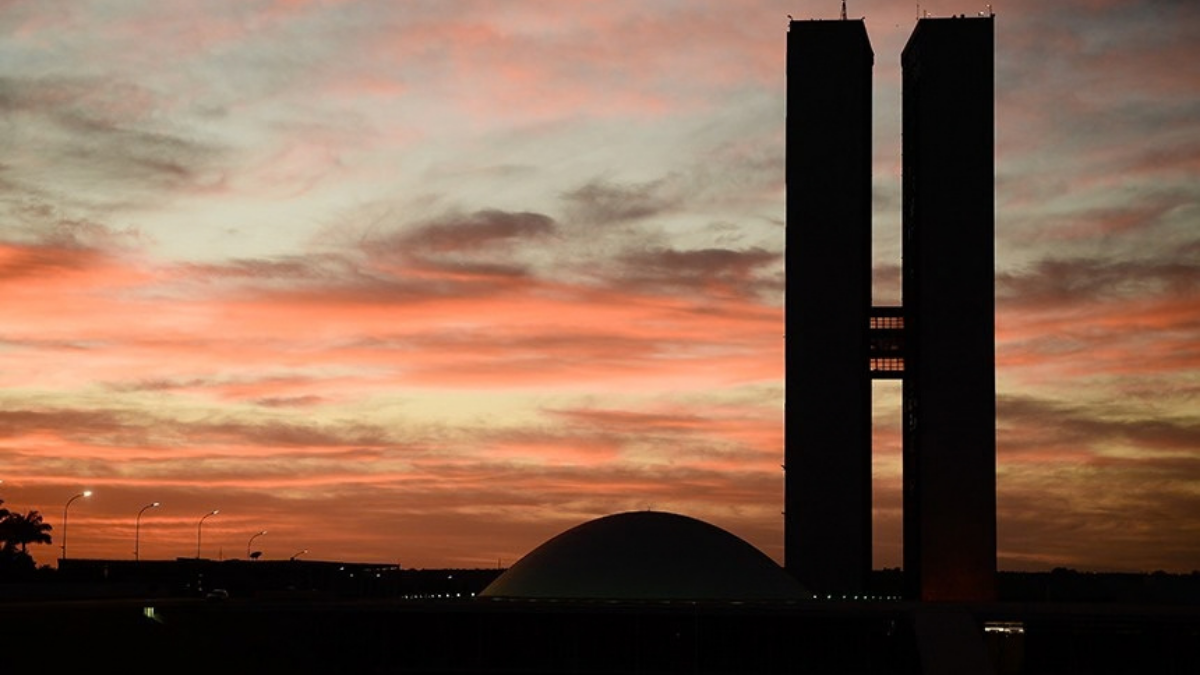 Silhueta do prédio do senado à beira de uma área aberta ao pôr do sol, com céu em tons laranja e nuvens ao fundo. Foto panorâmica ao entardecer.