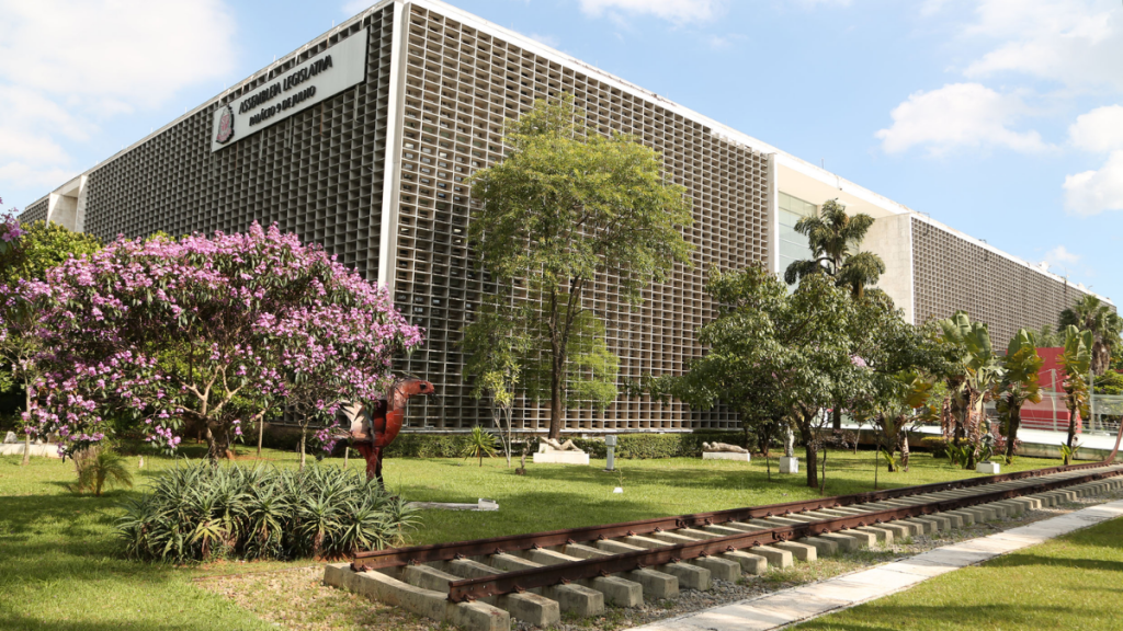 Prédio da Alesp em grelha com fachada de concreto e janelas quadriculadas, cercado por jardins bem cuidados, árvores e flores ao ar livre.