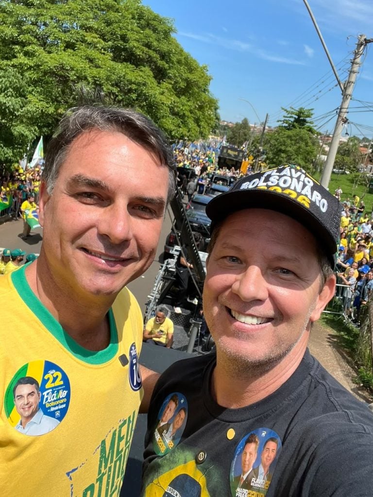 Flávio Bolsonaro e Mario Frias fazem selfie durante manifestação política, cercados por apoiadores vestidos de verde e amarelo.