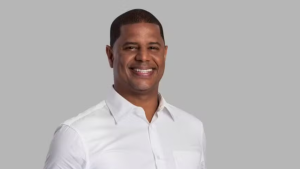 Marcelinho Carioca sorrindo, usando camisa social branca, em fundo cinza neutro, em foto corporativa de estúdio.