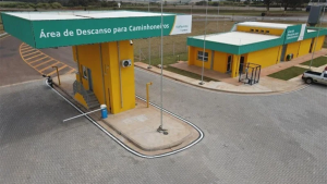 Vista aérea de uma área de descanso para caminhoneiros com duas estruturas amarelas e telhados verdes, acesso pavimentado e sinalização no pátio externo.