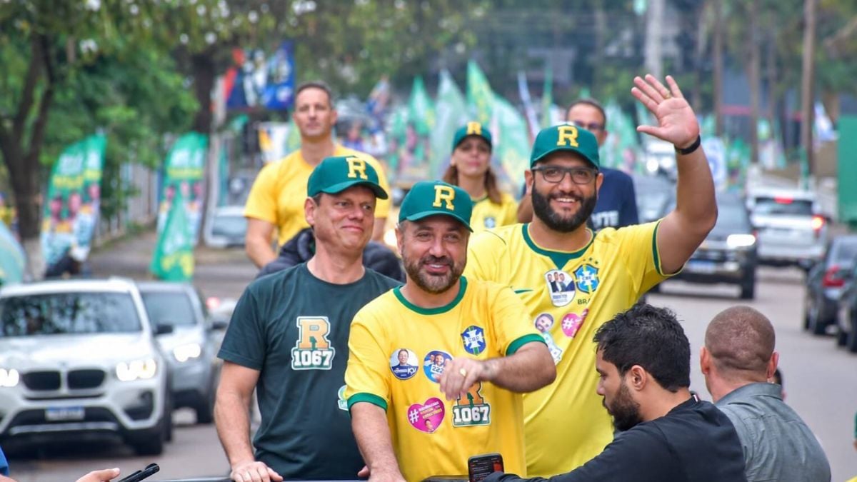 Campanha eleitoral em rua de cidade durante evento político, com homens usando camisetas e bonés verdes e amarelos com a letra R, um deles acenando para a câmera enquanto carros passam ao fundo Foto: Divulgação