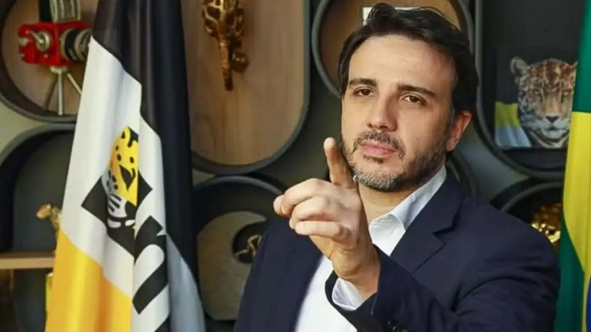 Renan Santos com terno azul e camisa social, em ambiente institucional, fazendo gesto de apontar com a mão, ao lado de bandeiras com símbolos e cores nacionais.