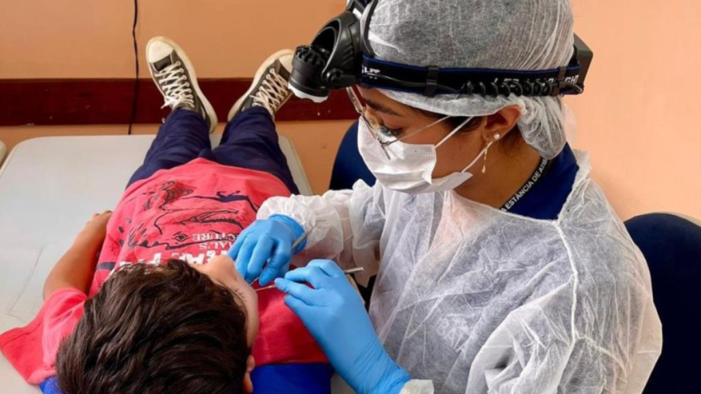 Profissional de saúde com máscara e viseira, usando luvas azuis, aplica cuidados em uma criança deitada, em ambiente clínico de vacinação ou atendimento médico.
