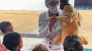 Profissional de saúde em ambiente externo usando máscara, segurando uma espécie de dentadura ou modelo odontológico amarelo e demonstrando a técnica para crianças em um momento educativo.