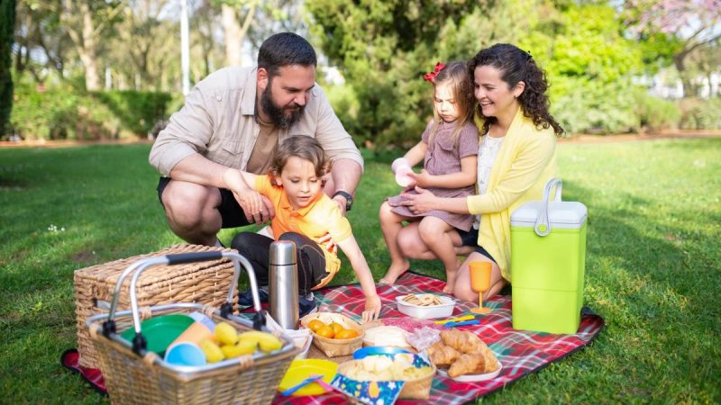 Família em um piquenique ao ar livre em dia de folga, com cesta e garrafa térmica no gramado enquanto pais brincam com as crianças no feriado