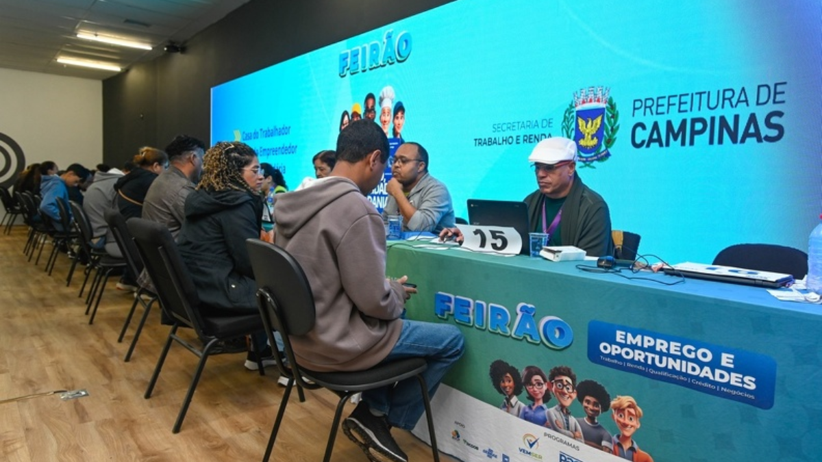 Mesa de atendimento em evento do Feirão, com participantes sentados e equipe em frente ao telão com a identificação “PREFEITURA DE CAMPINAS”, em Campinas.