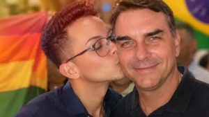 Foto de dois homens em destaque, João Pedro Sastre beijando a bochecha do Flávio Bolsonaro, em um clima de afeto, com bandeira colorida ao fundo.
