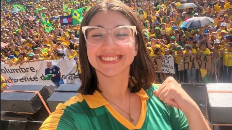 Eduarda Campopiano sorridente em meio a uma grande multidão durante evento ao ar livre, usando óculos e camiseta verde e amarela, com bandeiras ao fundo.
