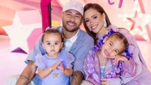 Neymar, Bruna Biancard e filhas sorridente em festa temática rosa e estrelas, com pai, mãe e duas crianças vestidas com acessórios coloridos, posando juntas para foto