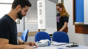 Funcionários em sala de votação e estão ao lado do equipamento e cadeiras azuis, com um banner de palestra ao fundo durante evento corporativo.