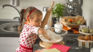 Dia das Crianças na cozinha: menina preparando massa com um rolo em bancada com tigela de farinha e ingredientes para receitas fáceis com os filhos.