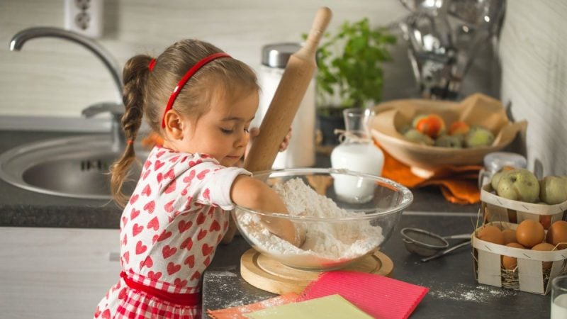 Dia das Crianças na cozinha: menina preparando massa com um rolo em bancada com tigela de farinha e ingredientes para receitas fáceis com os filhos.