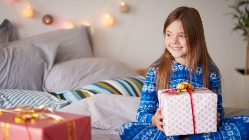 Criança sorridente em casa segurando um presente embrulhado em papel rosa com laço dourado, com luzes decorativas ao fundo. Dia das Crianças