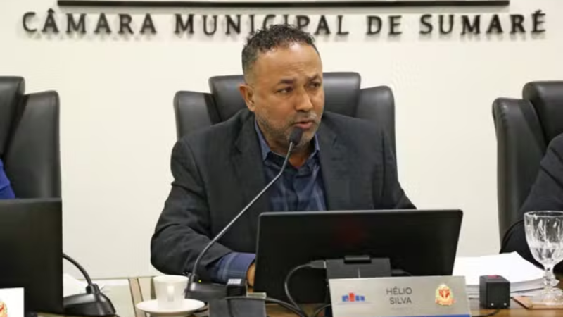 Reunião na CÂMARA MUNICIPAL DE SUMARÉ. Hélio Silva com terno fala ao microfone sentado à mesa, diante de um computador, em plenário.
