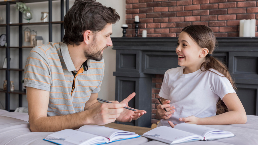 Pai e filha sentados à mesa conversando e gesticulando carinhosamente, com livros abertos sobre a superfície em um ambiente aconchegante com lareira decorativa e tijolos aparentes ao fundo.