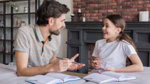 Pai e filha sentados à mesa conversando e gesticulando carinhosamente, com livros abertos sobre a superfície em um ambiente aconchegante com lareira decorativa e tijolos aparentes ao fundo.