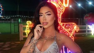 MC Mirella em ensaio noturno em quadra, segurando o rosto com a mão e usando biquíni prateado, com letreiro de luzes em formato de letras ao fundo