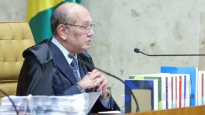 Ministro Gilmar Mendes em sessão solene no plenário, usando terno e óculos, com as mãos próximas ao peito diante de microfone, em uma mesa com pastas e bandeira ao fundo.