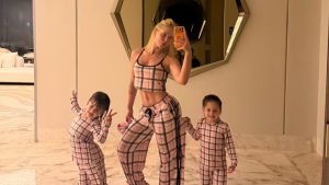 Virginia em frente ao espelho do banheiro, usando pijama xadrez rosa e branco, fazendo selfie com o celular; ao lado, suas duas filhas também em pijama xadrez.