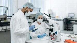 Pessoas em laboratório usam jalecos, máscaras e luvas de proteção enquanto realizam exame com amostras e instrumentos científicos, em ambiente de pesquisa, ilustrando informação sobre peste pneumônica, seus sintomas e risco