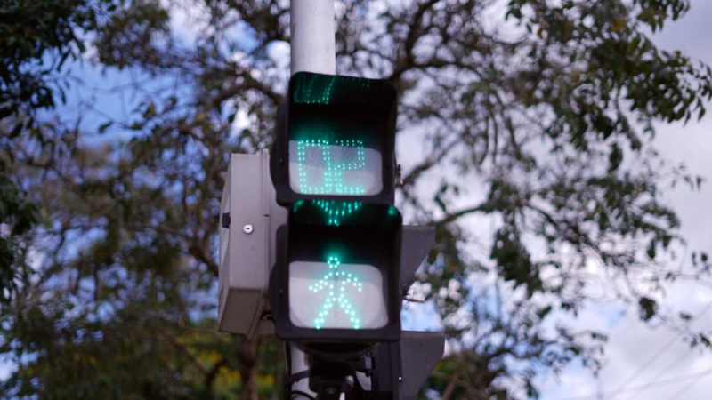 Semáforo de trânsito com sinal de pedestres verde e vermelho em funcionamento, com luzes LED visíveis ao fundo de árvores e céu nublado.