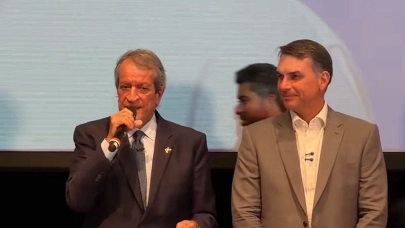 Valdemar Costa Neto e Flávio Bolsonaro em traje social em um palco, durante um evento, com um deles falando ao microfone e o outro sorrindo ao lado. Ao fundo, uma tela de projeção e pessoas em destaque ao menos ao redor.