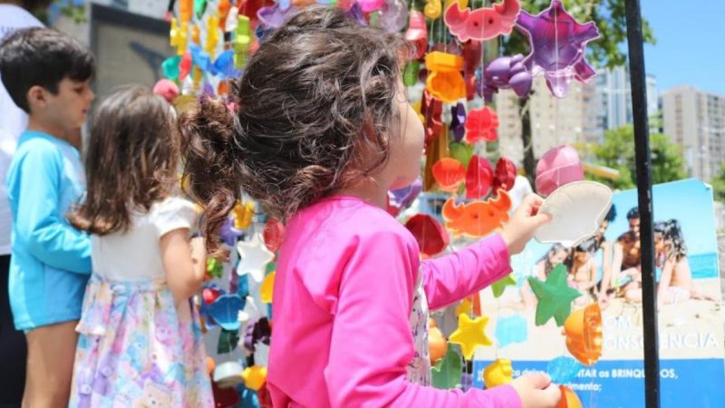Criança de costas com cabelo castanho encaracolado brincando em um estande decorado com balões e estrelas coloridas, em evento ao ar livre com outras pessoas ao fundo.