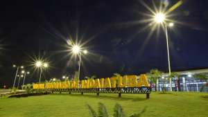 Placa com o nome “INDIATUBA” e gramado em primeiro plano, em uma área externa iluminada por postes de luz sob o céu noturno, com estrutura ao fundo.