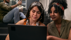 Duas mulheres em um ambiente de escritório, analisando informações no notebook. Elas usam faixas estampadas no cabelo e parecem concentradas durante uma conversa tranquila.